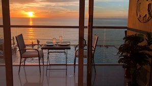 Un balcón con una mesa dispuesta para dos personas, con vista a una puesta de sol sobre el mar.
