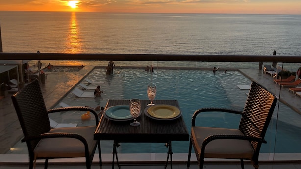 Vista de la piscina al atardecer sobre el océano, con sillones de descanso y una mesa dispuesta para dos.