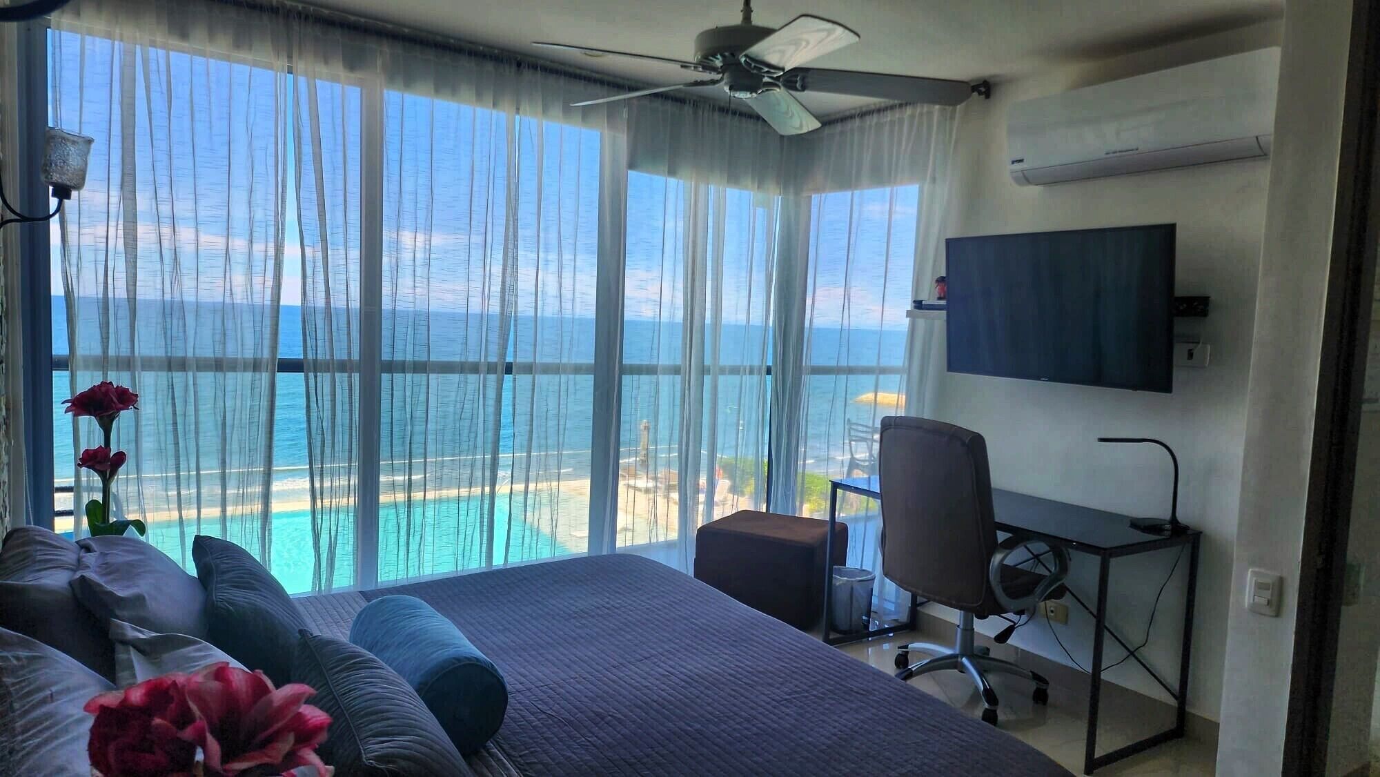 A hotel room with a bed, a desk, a chair, and a TV. The room has a large window with a view of the ocean.