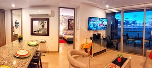 A modern living room with a kitchen area, a dining table with plates, a TV mounted on the wall, and a view of the ocean through large windows. - Morroscity - Amazing Poolside Paradise, Ultra Fast Internet, 1BR 2Bath, 5 Stars (Cartagena)