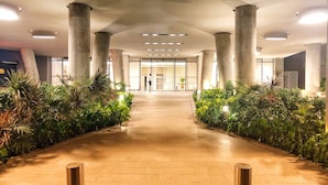 Un pasillo bien iluminado con un camino central flanqueado por columnas y abundante vegetación, que conduce a la entrada de un edificio.