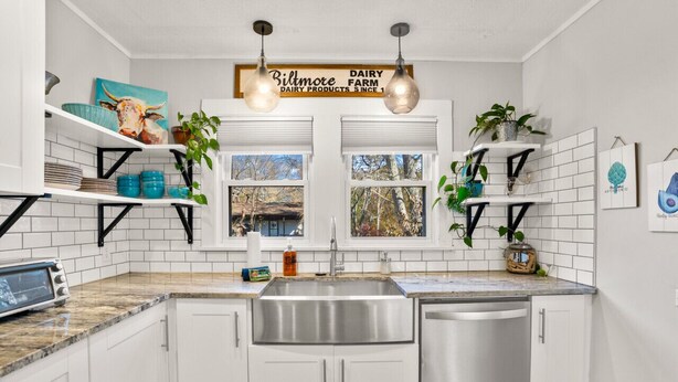 A modern kitchen with white cabinets, a stainless steel sink, and a dishwasher. There are two windows with views of trees and a barn. Shelves hold kitchenware and plants. - Fun Rec room, Hot Tub & Mountain View’s 2 King Rooms (Candler)