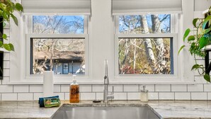 A kitchen with a stainless steel sink, two windows with views of trees and a house, and a countertop with a bottle of hand soap and a paper towel dispenser. - Fun Rec room, Hot Tub & Mountain View’s 2 King Rooms (Candler)