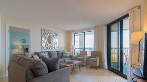 Een woonkamer met een grijze bank, twee fauteuils, een salontafel en een grote spiegel. Er is een balkon met uitzicht op zee.