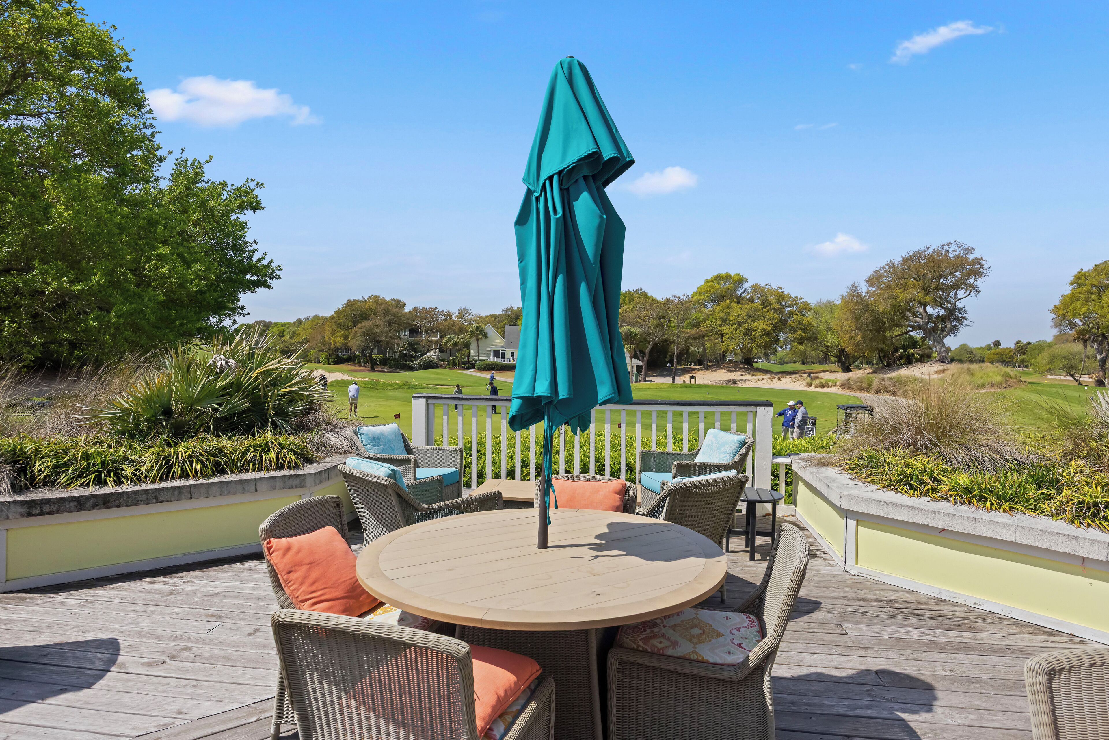 Een ronde houten tafel met rieten stoelen en een blauwgroene parasol op een houten terras met uitzicht op een golfbaan.