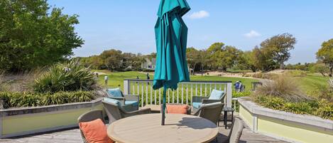 Een ronde houten tafel met rieten stoelen en een blauwgroene parasol op een houten terras met uitzicht op een golfbaan.