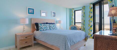 A bedroom with a bed, two bedside tables, a chair, and a view of the beach.