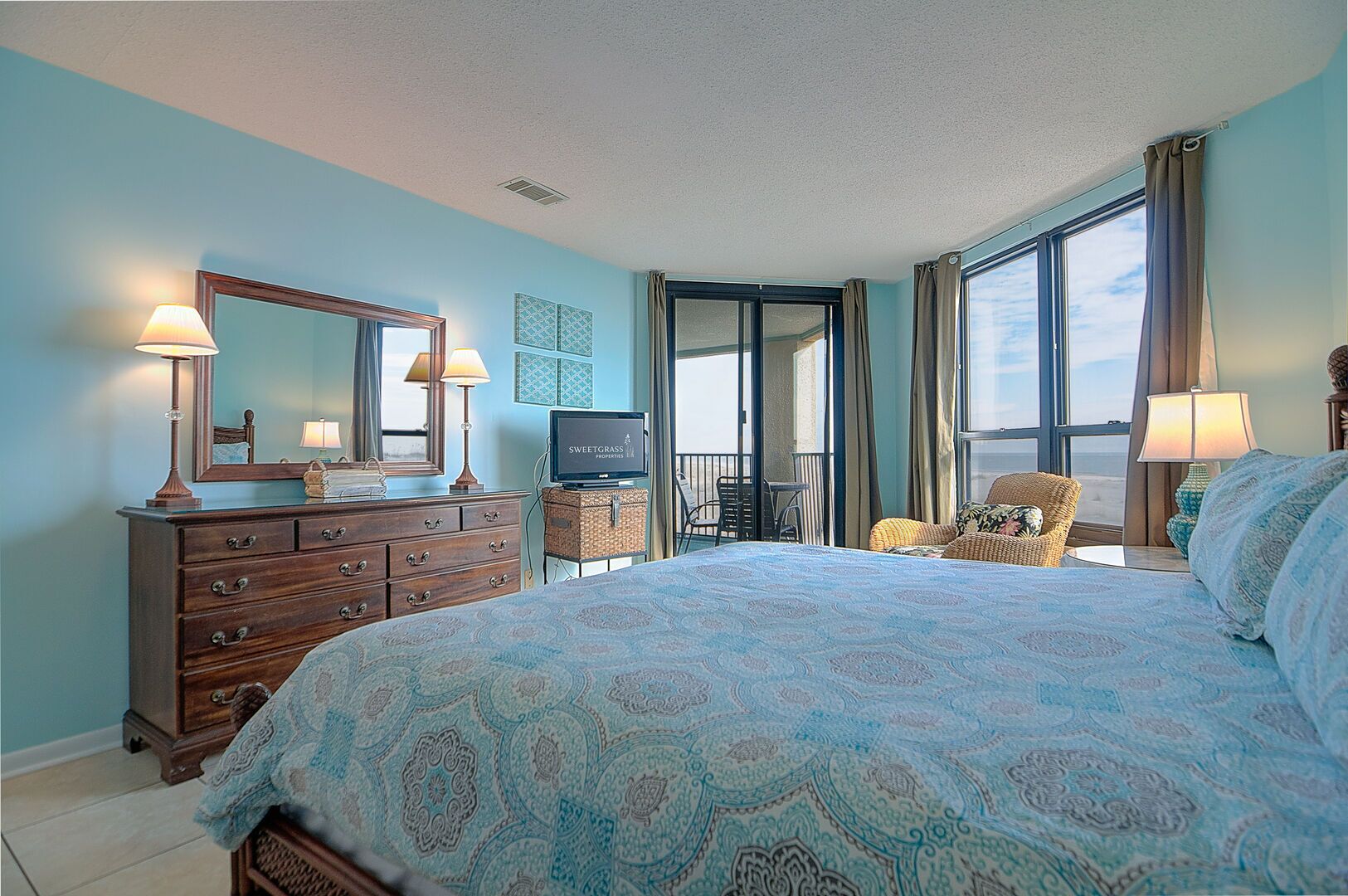 A bedroom with a large bed, a dresser, a television, and a balcony with a chair.