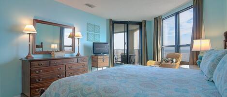 A bedroom with a large bed, a dresser, a television, and a balcony with a chair.