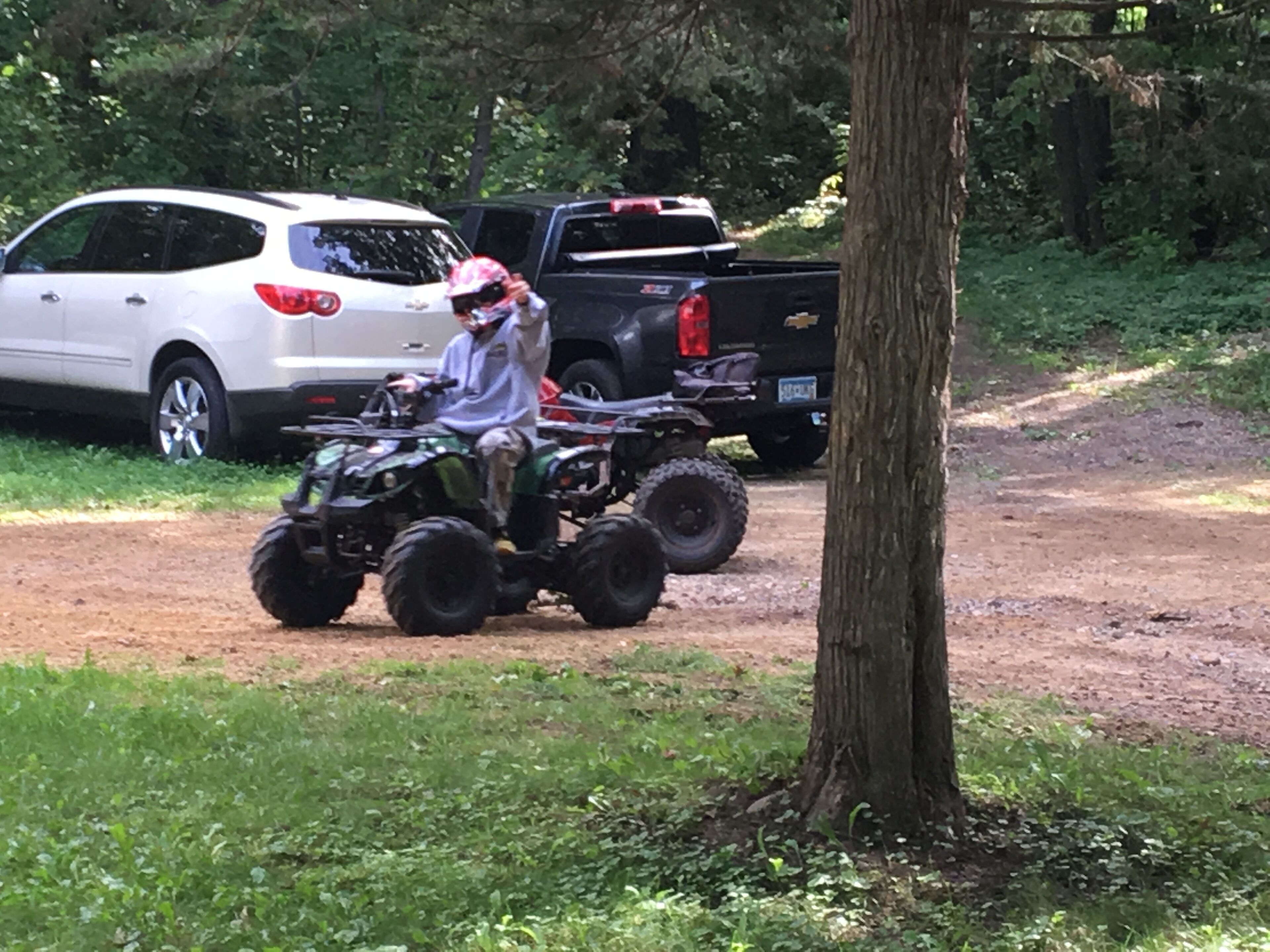 En person kører på en ATV på en grusvej, mens en hvid SUV og en sort pickup er parkeret i nærheden.