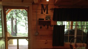 A cabin interior with a wooden door leading outside, a wall-mounted 'M' sign, a welcome sign, and a dining area with chairs and a table. - Cozy Authentic Amish Cabin, 5 Acres Of Privacy, 4 Wheel Trail, Snowmobile Trail (Lampson)