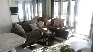 Ein Wohnzimmer mit einem Ecksofa, einem Glastisch und einem kleinen Beistelltisch mit einer Kerze.