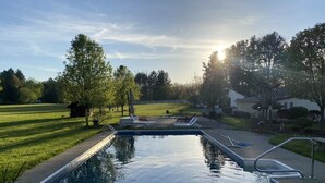 Une piscine rectangulaire avec un escalier en métal et un petit espace aménagé autour, entourée d’une pelouse verdoyante et d’arbres.