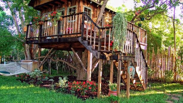A wooden treehouse with multiple levels, surrounded by lush greenery and flowering plants. - Rural appeal yet so close to town (Taos)