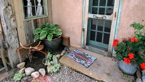A rustic building with a wooden door, a window with curtains, and a small garden area with potted plants and a colorful mat. - Rural appeal yet so close to town (Taos)