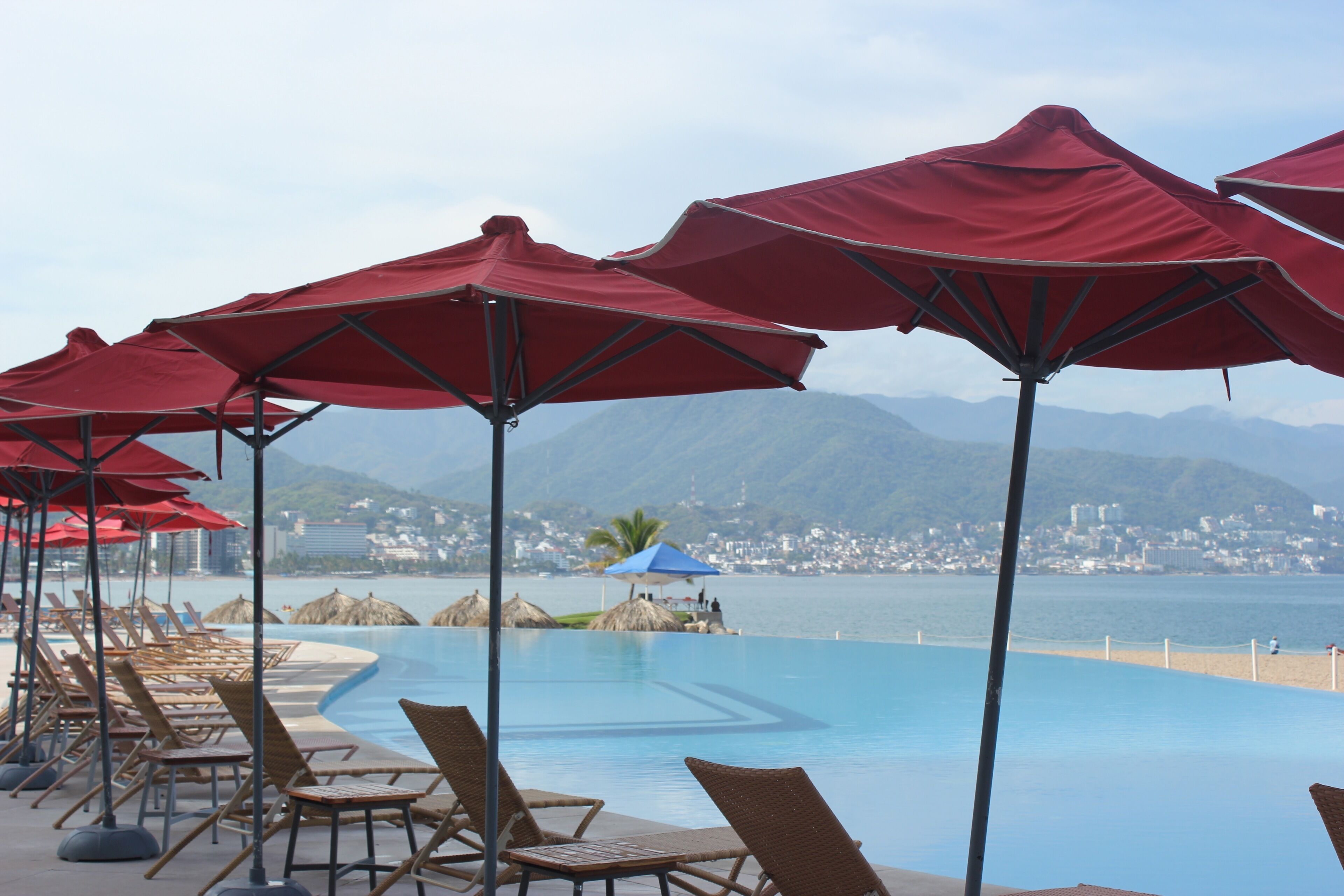 Area piscina con ombrelloni rossi, lettini e vista sul mare e sulle montagne.