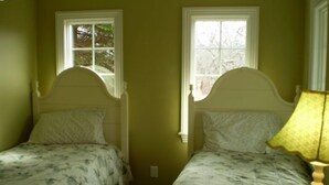 A room with two single beds, each with a white headboard and floral bedding. There is a window on the left side of the room, and a lamp on the right side. - Charming Chappaquiddick Retreat: Steps from Private Beach & Edgartown's Best! (Edgartown)