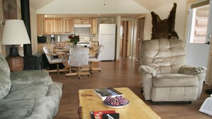 Ein geräumiger Wohnbereich mit Sofa, Sessel und Couchtisch. Im Hintergrund ist eine Küche mit Holzschränken und einem Kühlschrank zu sehen.