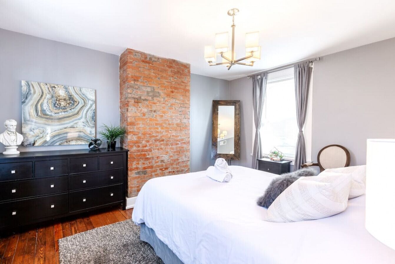 A modern bedroom with a brick accent wall, a large bed with white linens, a black dresser, and a chandelier.