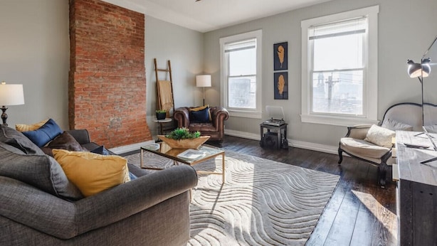 A spacious living room with a brick wall, leather sofa, and a large window. - OTR Penthouse: Walk Everywhere & FREE Parking (Cincinnati)