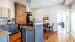 A modern kitchen with a brick accent wall, a stainless steel range hood, and a dining area with a table and chairs. - OTR Penthouse: Walk Everywhere & FREE Parking (Cincinnati)