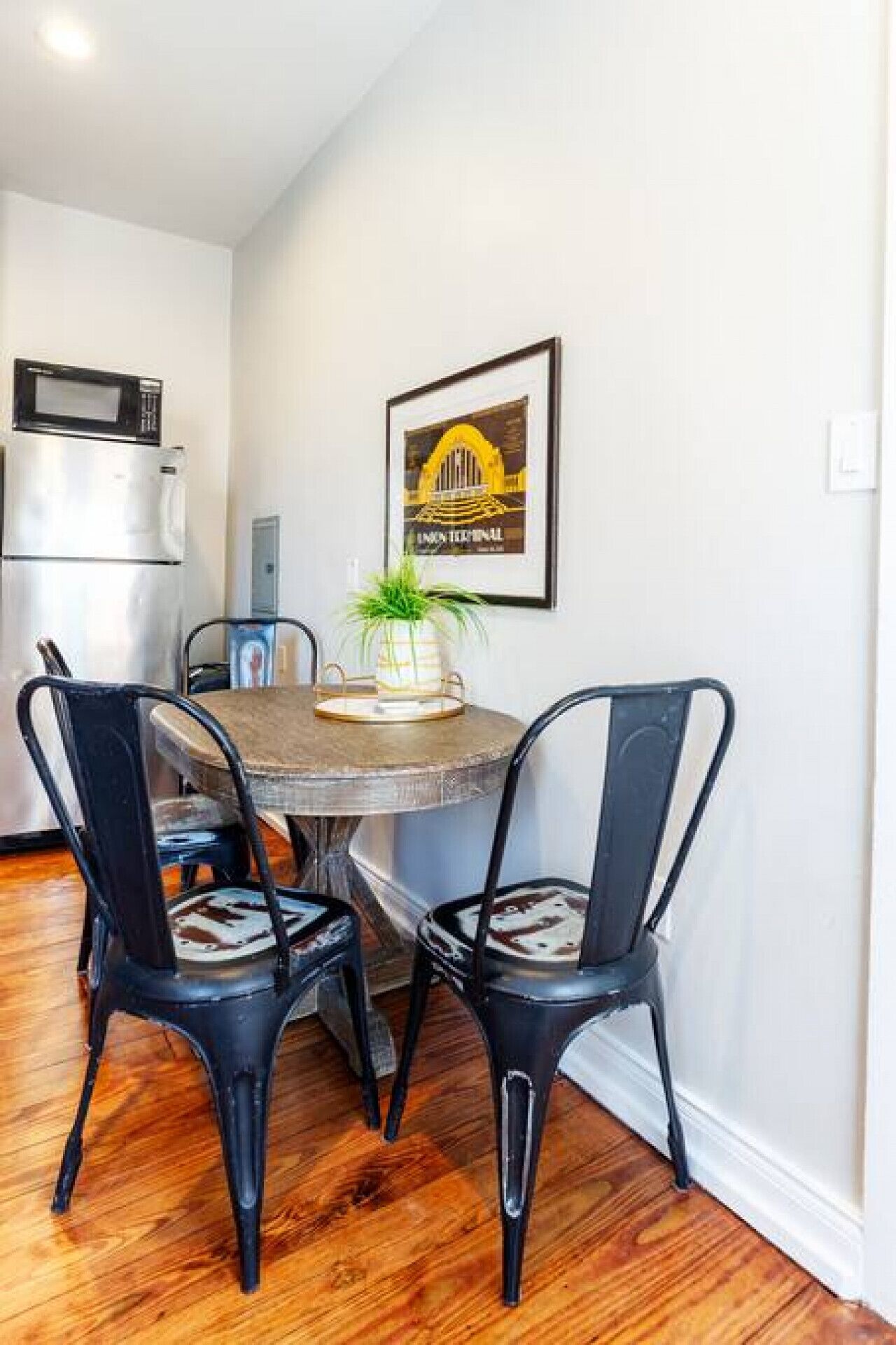 A dining area with a round table, two chairs, a potted plant, a framed picture, and a microwave above the table.
