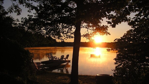 A lakeside scene with a dock, chairs, and a bench under a tree during sunset.