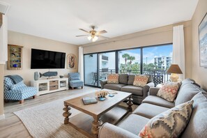 A spacious living room with a large TV, comfortable seating, a ceiling fan, and a balcony with a view of palm trees and buildings.