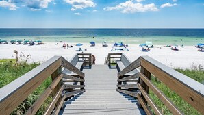 Eine Holztreppe führt zu einem Strand mit blauem Himmel und Sonnenschirmen.