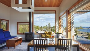 A modern living room with a wooden ceiling, large windows, a blue sofa, a dining table with chairs, and a view of the sea. - The Horizon- Award Winning- Sauna Available for a fee (Grand Marais)