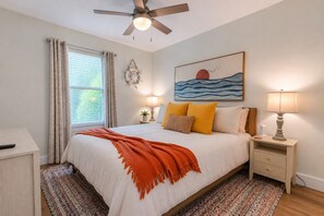 A bedroom with a bed, a ceiling fan, a nightstand with a lamp, a bedside table, a window with curtains, and a wall-mounted artwork.