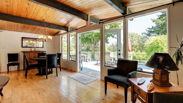 A spacious room with wooden flooring, a dining area with a table and chairs, a large window with a view of greenery, and a ceiling with exposed beams. - A lakefront playground near beautiful Victoria on British Columbia's west coast. (victoria)