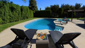 Un patio trasero con piscina, dos sillones de descanso y una mesa con bebidas y aperitivos.