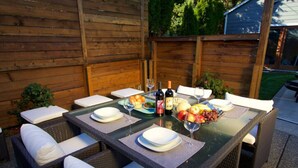Un área de comedor al aire libre con una mesa de vidrio preparada para cuatro personas, rodeada de sillas de mimbre con cojines blancos.