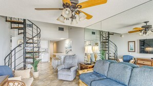 A spacious living area with a large blue sofa, a grey armchair, and a wicker chair. A spiral staircase leads to an upper level. - Spectacular 3 BR/3BA Oceanfront Condo -2 Large Balconies- Best Views-Aug wks ! (North Myrtle Beach)