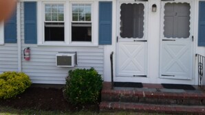 A single-story house with a white exterior, blue shutters, a front door with a decorative design, a small front yard with a bush, and a fire extinguisher mounted on the wall. - Dennisport - Studio Condo w kitchenette  - Walk to beach  (Dennis)