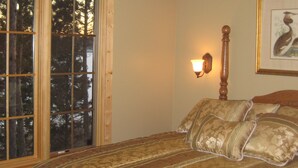 Ein Schlafzimmer mit großem Fenster, einem Bett mit gemusterten Bettwaren, einer Wandlampe und einem gerahmten Bild an der Wand.