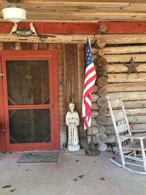 Entrada a una cabaña de madera con un letrero que dice 'Welcome', una estatua, una bandera estadounidense y una mecedora.