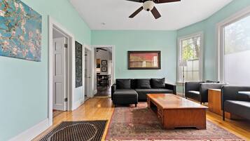 A spacious living room with a ceiling fan, a black sofa, a wooden coffee table, and a large window providing natural light.