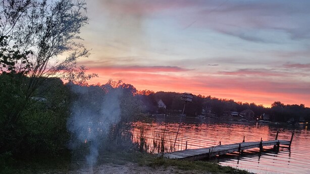 A campfire on a sandy beach with a wooden dock and chairs, overlooking a calm lake with a sunset in the background. -  the "LAKE HOUSE" - PONTOON INCLUDED (Middleville)
