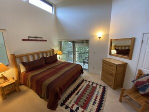 Une chambre avec un grand lit, une commode en bois, une table de chevet, un miroir et un balcon donnant sur un paysage verdoyant.