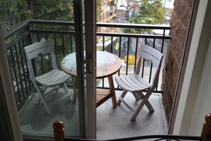 Un balcon avec deux chaises blanches et une petite table ronde, donnant sur une rue bordée d’arbres et de bâtiments.
