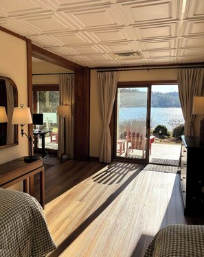 A hotel room with a wooden floor, a bed, a desk, a chair, and a view of a lake through large windows.