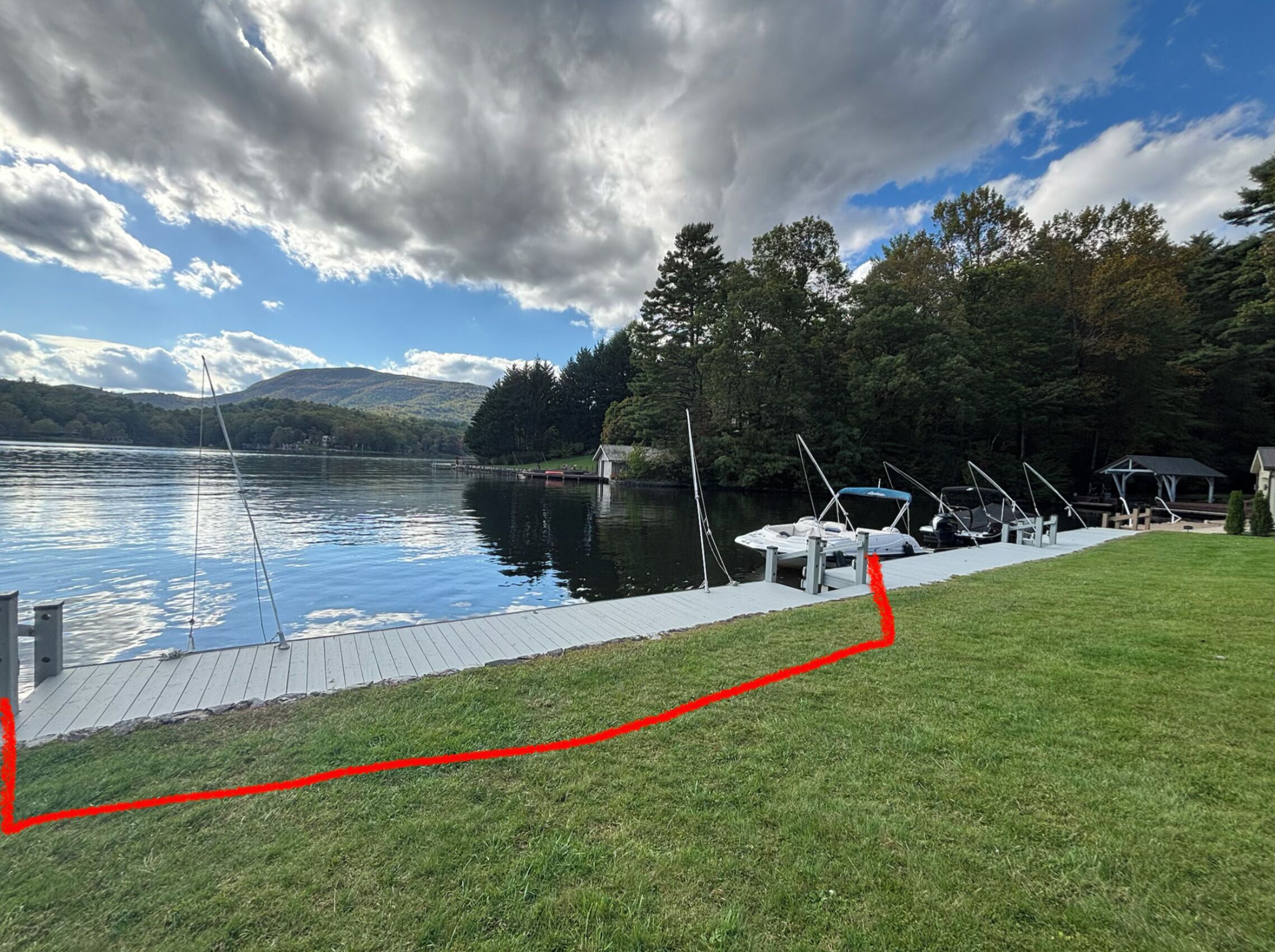 A lakeside dock with boats, a grassy area, and a forested hill in the background.