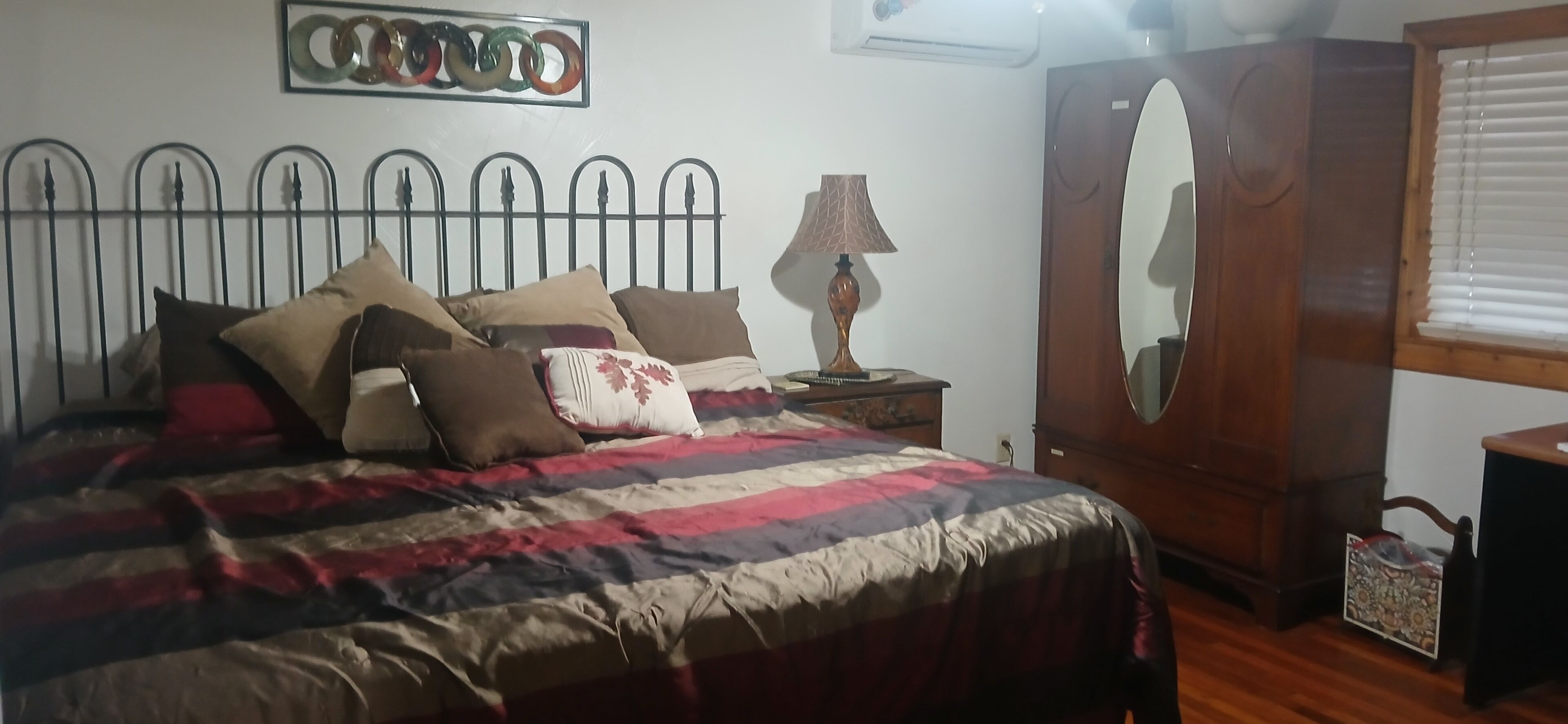 Quarto com estrutura de cama de metal, colcha estampada, abajur, guarda-roupa de madeira e espelho.