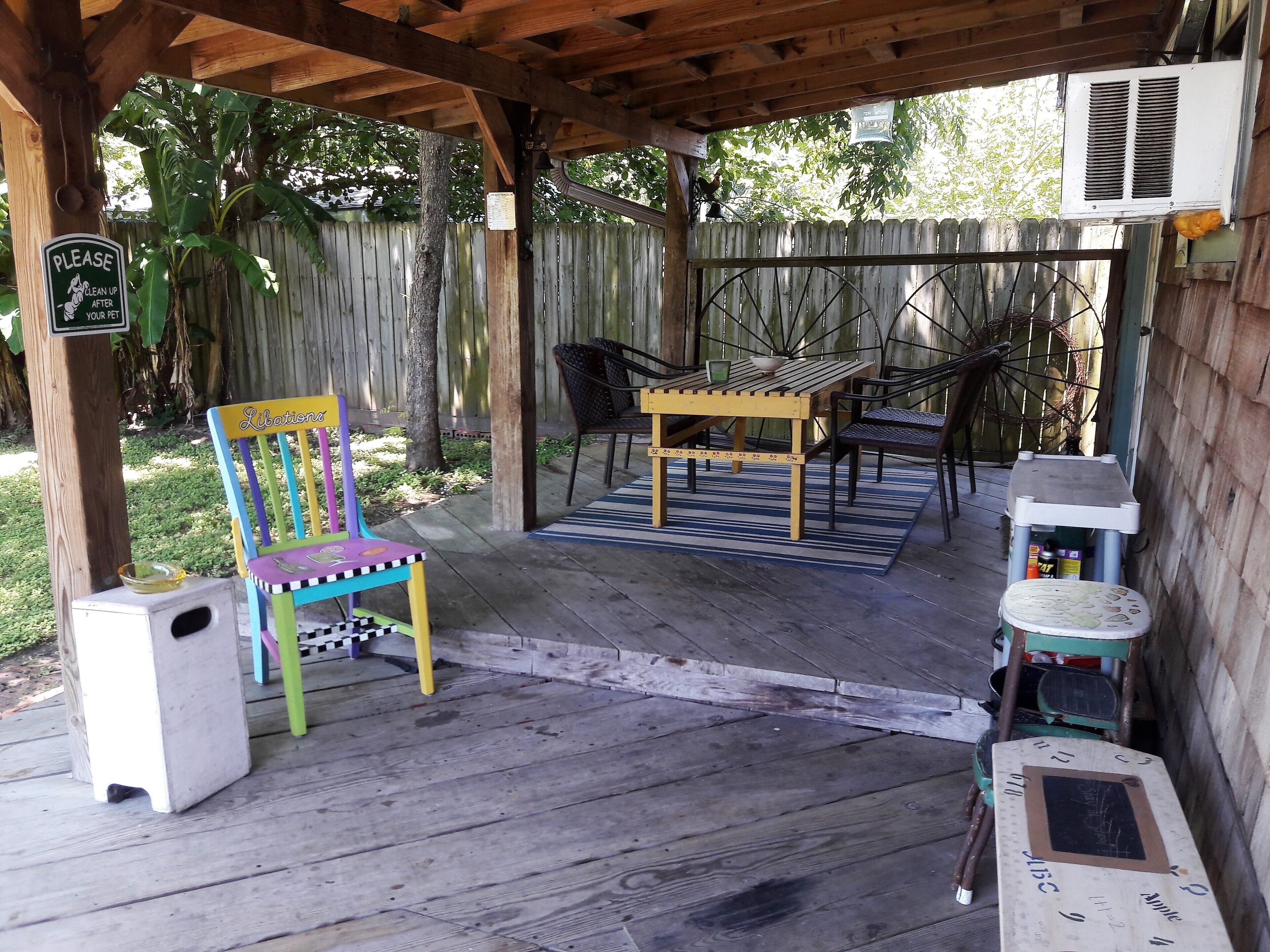 A covered patio area with a wooden table and chairs, a colorful chair, a small trash bin, and a white cooler.
