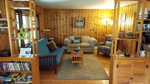 A cozy living room with wooden walls, a blue sofa, a beige armchair, and a small coffee table. There are bookshelves filled with books and a lamp on a side table.