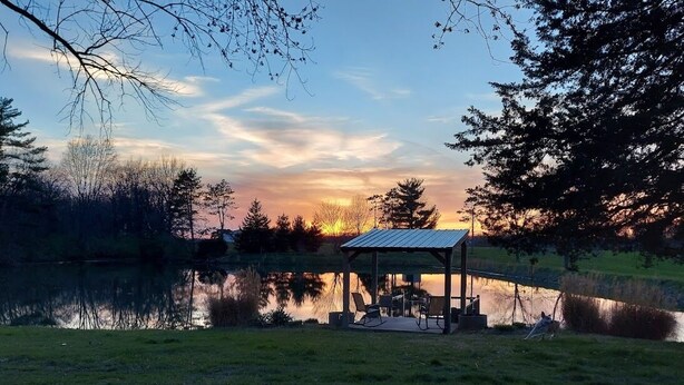 A gazebo with a metal roof and seating area by a calm lake at sunset.