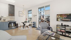 A modern living room with a grey sofa, a coffee table with books, a bookshelf, and a large window with a view of trees. - Sunny Chic Baca Railyard Retreat: Balcony + Full Kitchen + Laundry (Santa Fe)