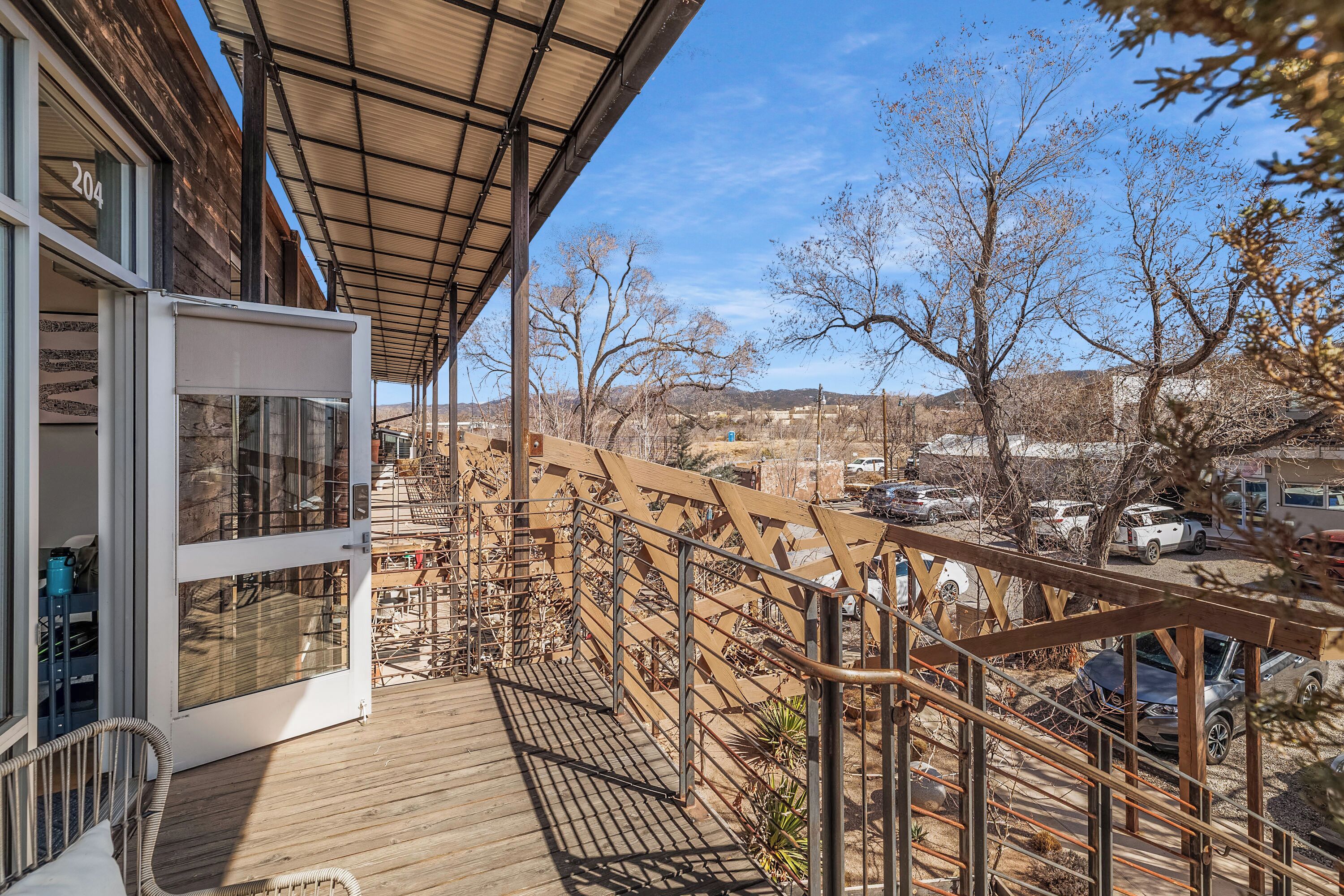 A wooden deck with a glass door leading to a room, a metal railing, and a view of a parking area and trees.
